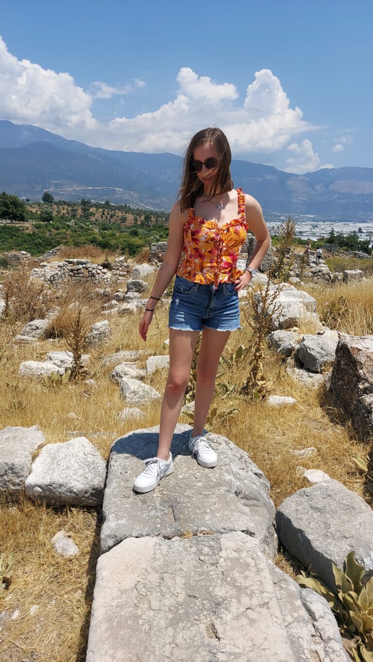 Image of Becky on holiday wearing a orange and red flowery top, denim shorts and sunglasses, posing on a giant rock on a mountain, smiling and looking down