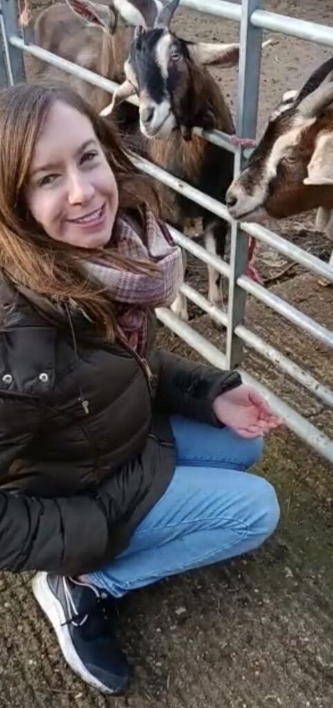 Photo taken of Becky kneeling down smiling, with goats poking their heads through the gate.