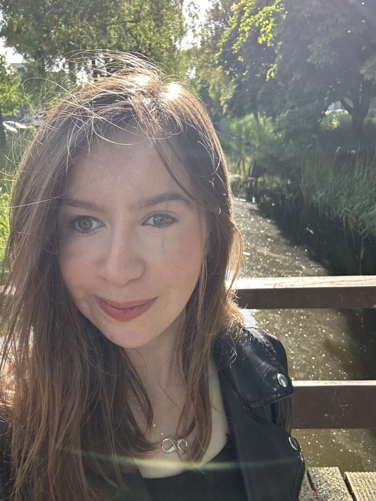 Selfie of Becky on a bridge over a stream in Amsterdam. Becky has mid length brown hair and is wearing a black top with puffed sleeves.